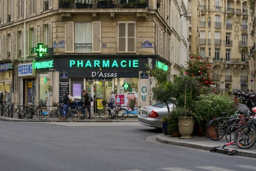 The exterior of Pharmacie D'Assas, a Parisian pharmacy on a street corner, with its name displayed in green neon lettering above the shopfront, bicycles parked outside and a decorated Christmas tree nearby.