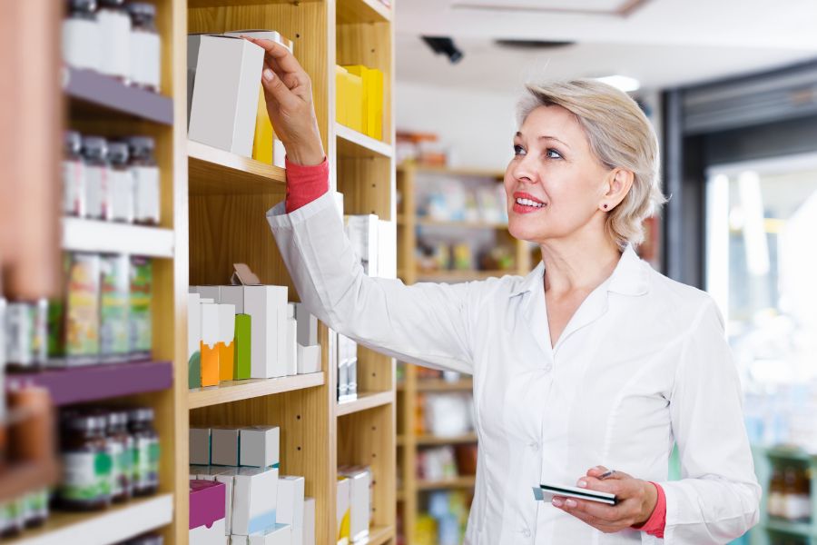 A smiling female pharmacist in a white lab coat reaches for a box on a wooden shelf stocked with various packaged products, holding a notepad in her other hand.
