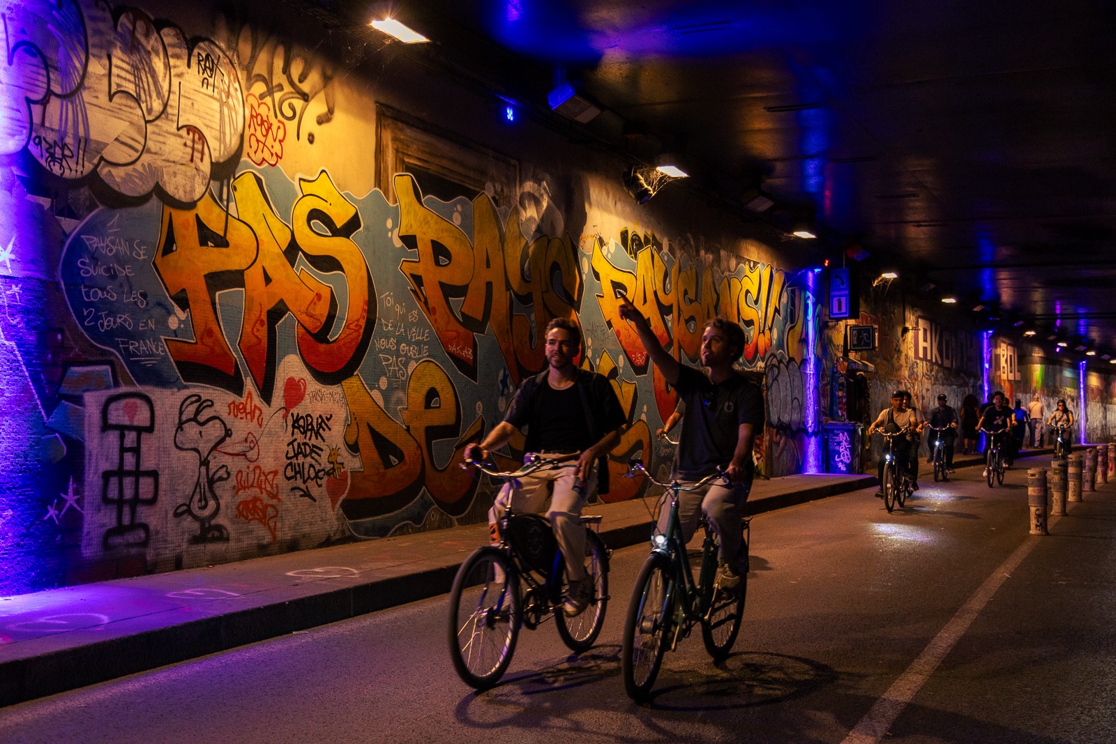 A group of young people riding bikes along a street art lined road in Paris, during the evening.