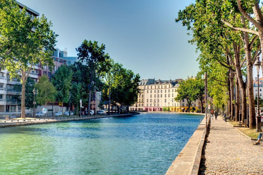 This is an image of a Paris canal on a sunny day with trees and buildings lining it.