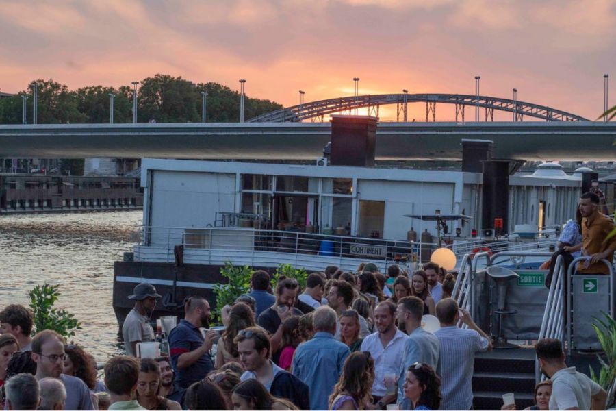 This is a picture of an open-air bar by the water with lots of people drinking and dancing together. There is a mild sunset behind it.
