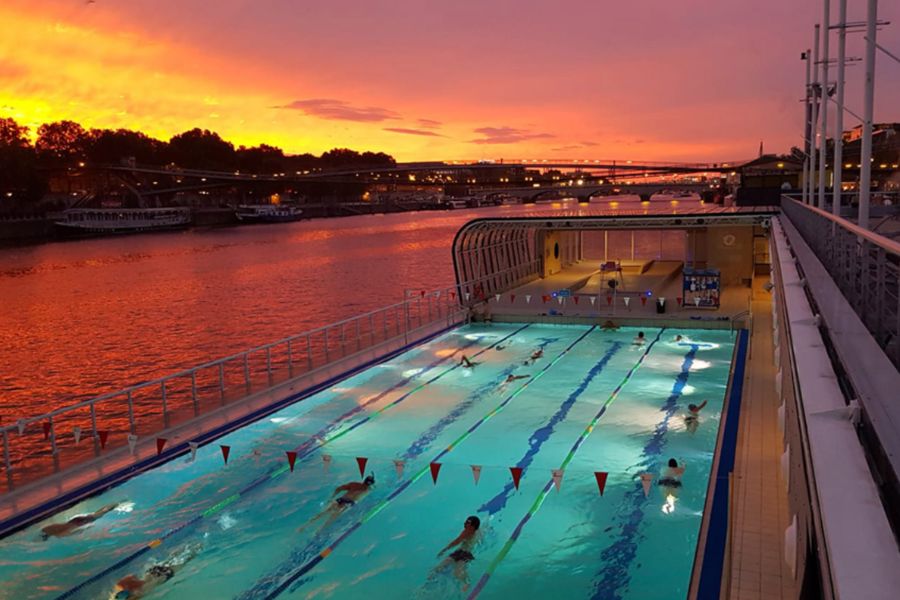 This is an image of an outdoor swimming pool along the Seine. There is a beautiful deep orange sunset behind it.