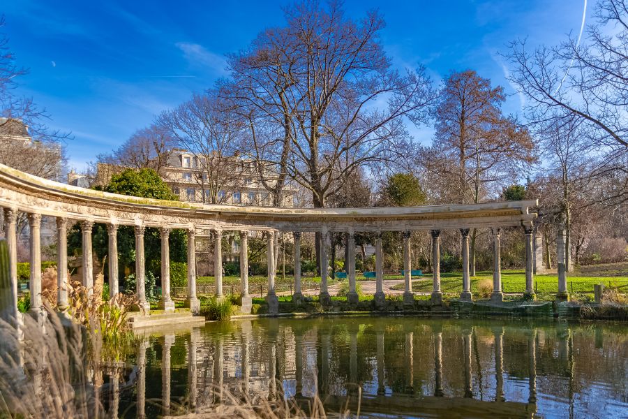 This is an image of a historic park called Parc Monceau. There is a small pond in the centre and some historic architecture surrounding it. The trees and leaves are green.