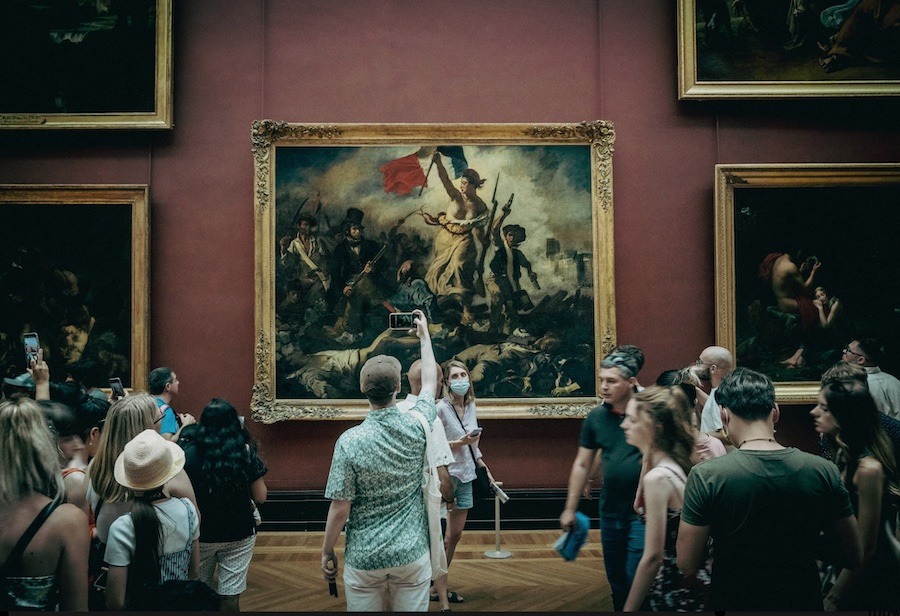 This is an image of a group of people stood around an old masters painting in an art gallery. Some people are taking photographs with their phones and some are admiring the painting whilst others are walking around the room. The louvre is such a famous museums, but this tour really is one of the most Unique Tours To Take While Visiting Paris.