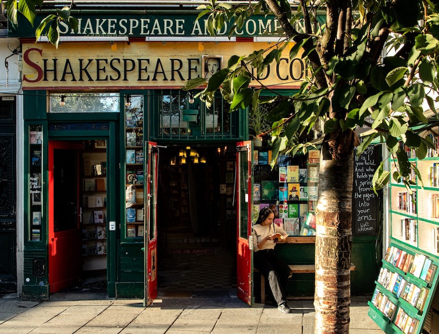 This is an image of a storefront that reads Shakespeare and Company on its front. The writing is old fashioned and it looks dimly lit inside the shop. There are various books displayed outside the store and there is also a tree in the foreground of the image covering up some of the writing of the stores name.