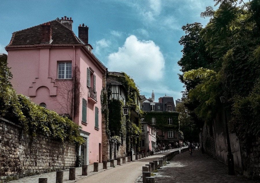 This is an image of a street in Montmatre, Paris. It is whimsical with a pink hour and lots of bushes overgrown on the houses. The sky is bright and blue in the background and it is a pleasing image. This is one of the most Unique Tours To Take While Visiting Paris.