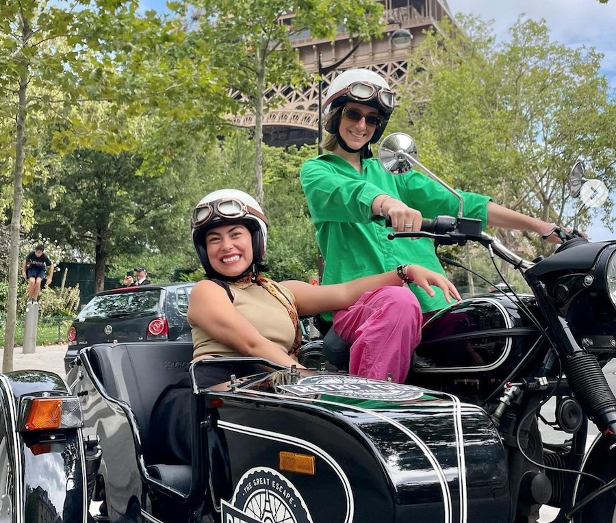 This is an image of two people in a vintage sidecar smiling and looking happy. The Eiffel Tower is in the background and trees as well. This is one of the most Unique Tours To Take While Visiting Paris.