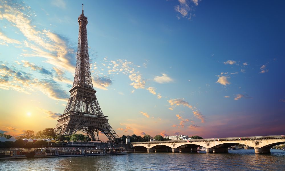 This is an image of the Eiffel Tower from across the Seine, as well as a beautiful arched bridge. There are clouds and a purple sunset in the back.