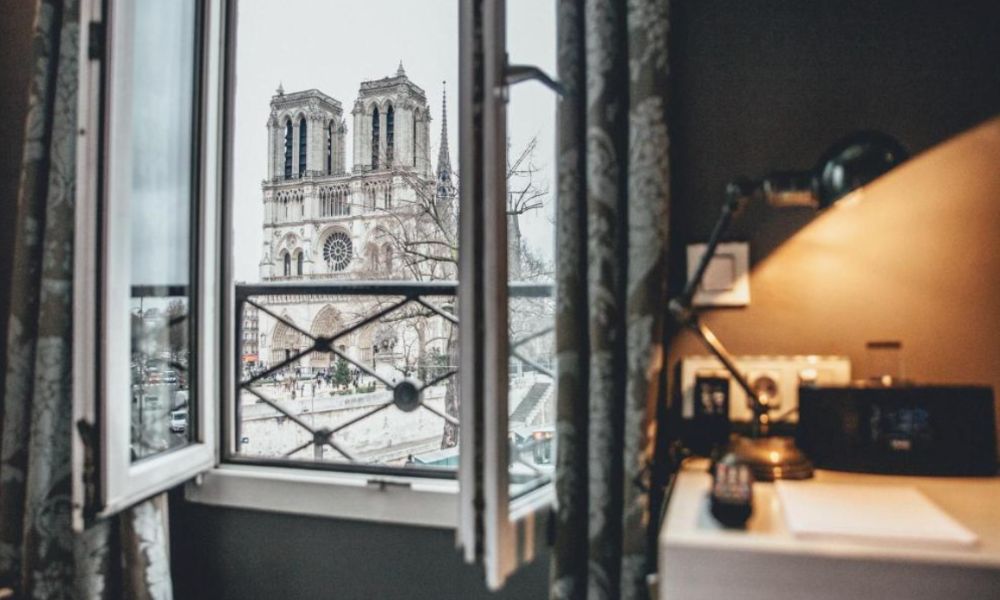This is an image of a dark, hotel room with a lamp and a window with the Notre Dame outside.