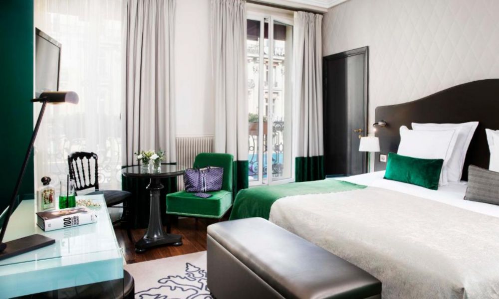 This is an image of a large hotel room with a big bed, vanity and large window with blinds. The bedding and decor is all white, and has jade-coloured accents.