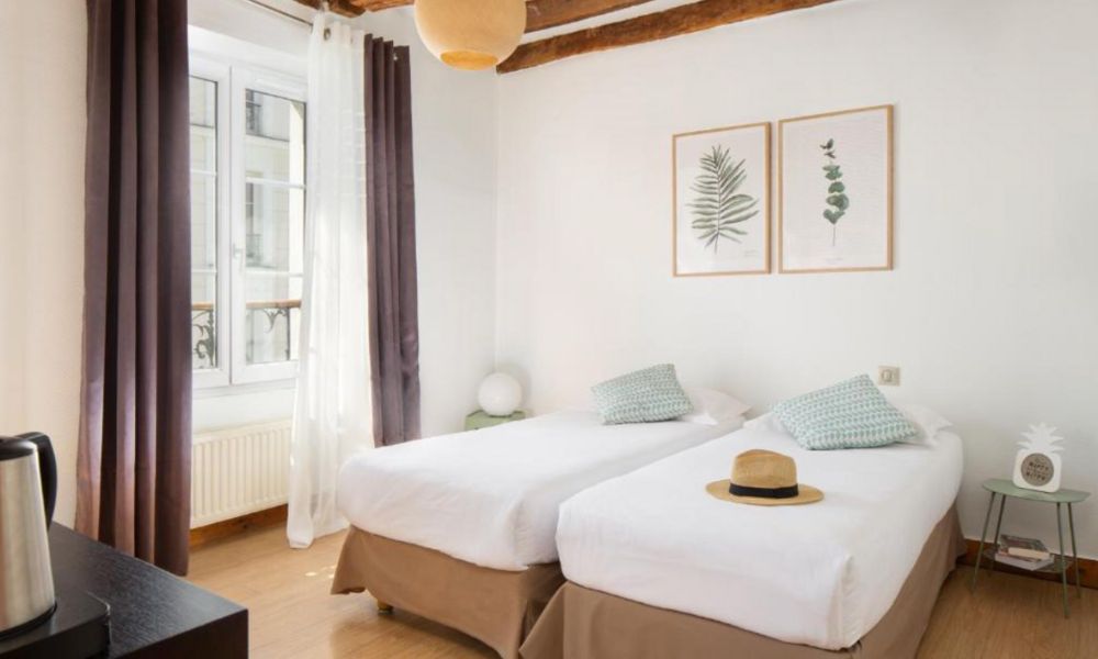This is an image of a very minimalist white room with two single beds in the middle. You can see a small table with a kettle and some paintings on the wall.