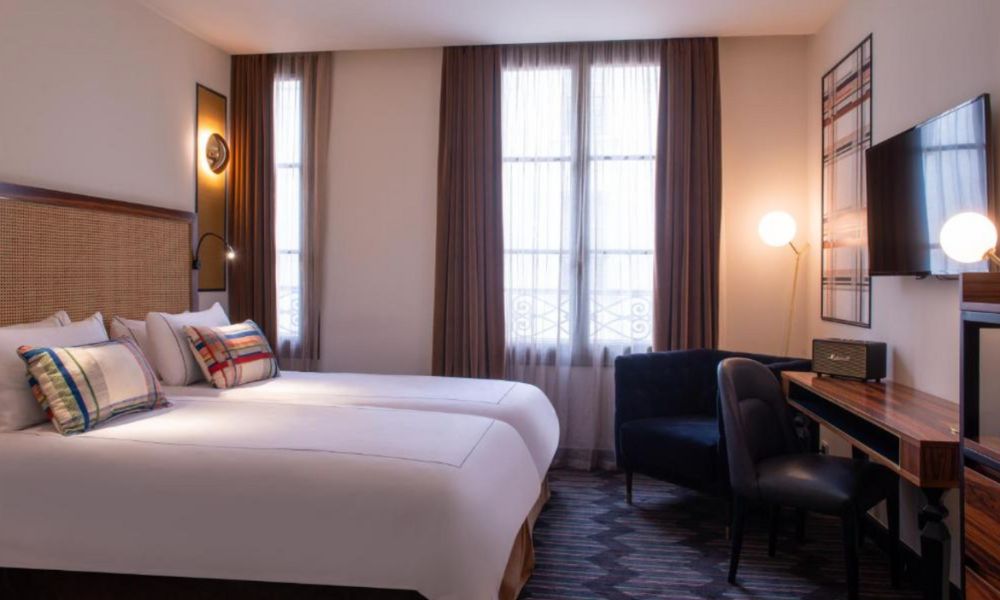 This is an image of a chic hotel room with artsy pillows on each of the single beds and cool artwork in the corner of the room. There is also a big window with curtains.