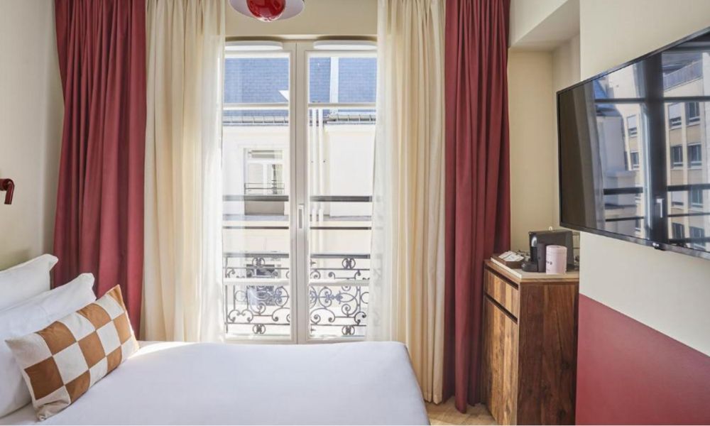 This is an image of a really minimalist, cream hotel room with a double bed and a TV on the wall opposite it. The curtains are pink and there are other pink accents around the room.