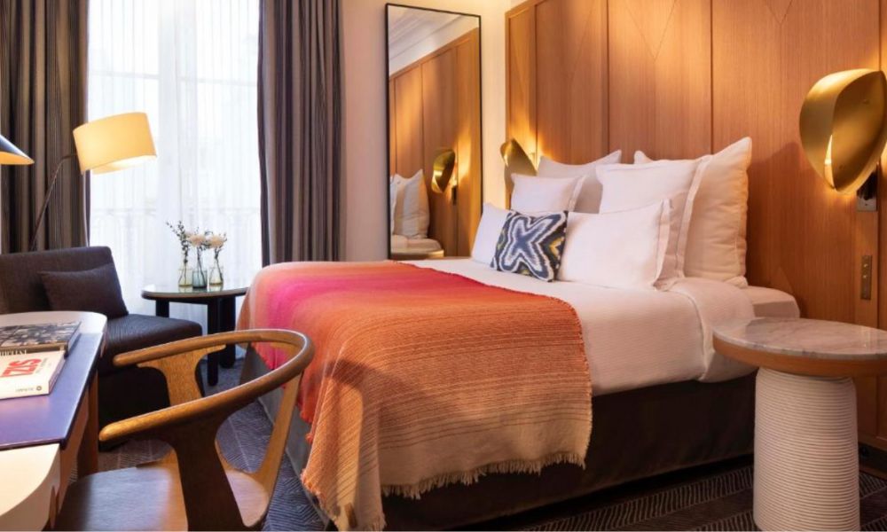 This is an image of a art hotel with a big bed, desk, lounger and coffee table, plus big gold lighting. There is also a big open window with blinds.