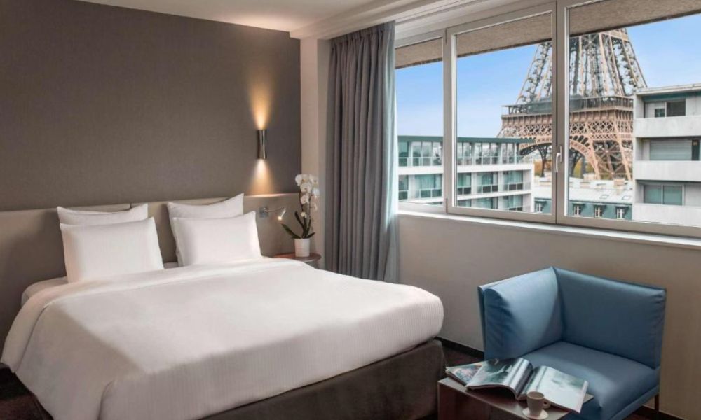 This is an image of a simple hotel room with a double bed, a chair and coffee table, plus a big window with a view of the Eiffel Tower really close.