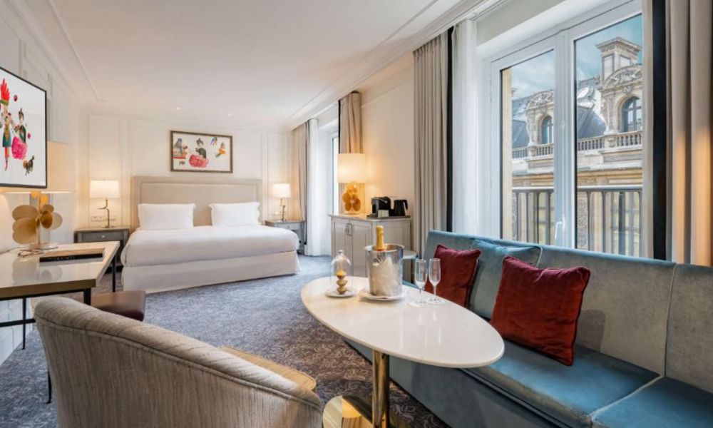 This is an image of a large hotel room with a queen bed, plus a lounge area and large awning windows.