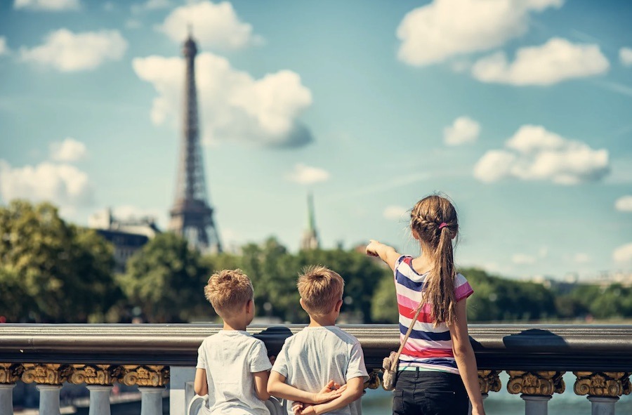This is an image of three kids on a bridge. The older girl is pointing up to the Eiffel Tower in the distance.