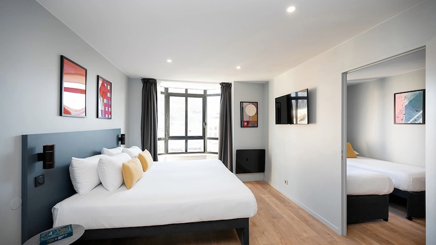 This is an image of a big hotel bedroom with a large double bed with white bedding and big windows that are letting in a lot of natural daylight. It is a minimalistic room.