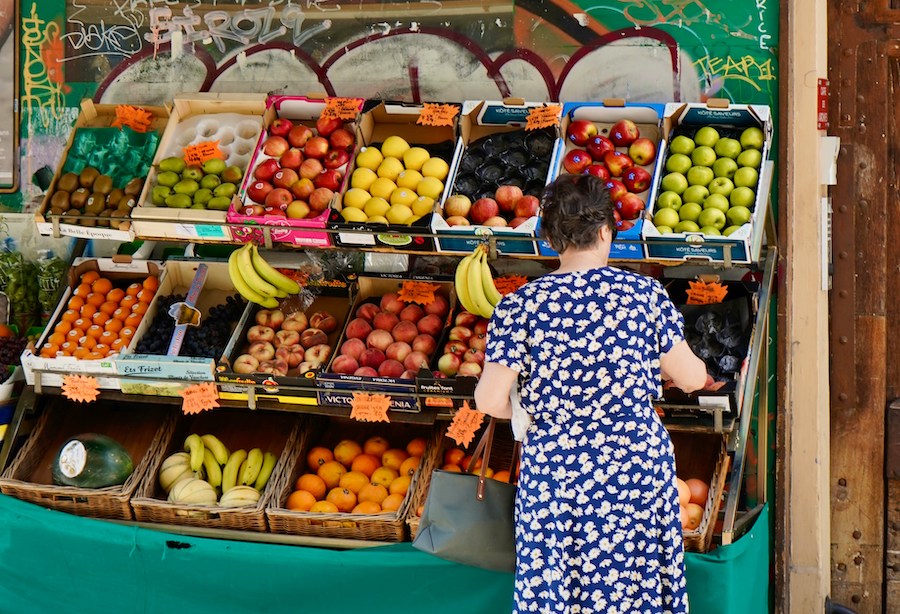 This is an image of a lady shopping at a local fresh food market. She is browsing the fruits, holding a bag of her purchases. This is one of the top Paris tours for food lovers.