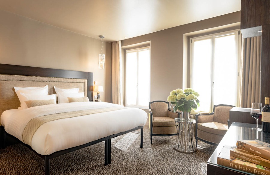 This is an image of a spacious hotel bedroom with a big double bed, muted coloured furniture and big windows that are letting in lots of natural daylight into the room.
