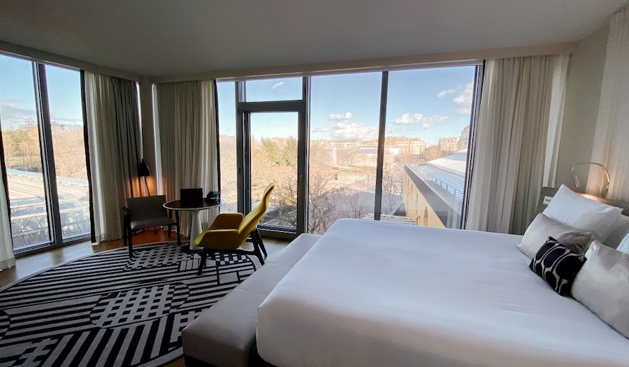 This is an image of a luxurious hotel bedroom with a big double bed, ceiling length windows with a view of Paris and modern furniture that is sleek and cool.