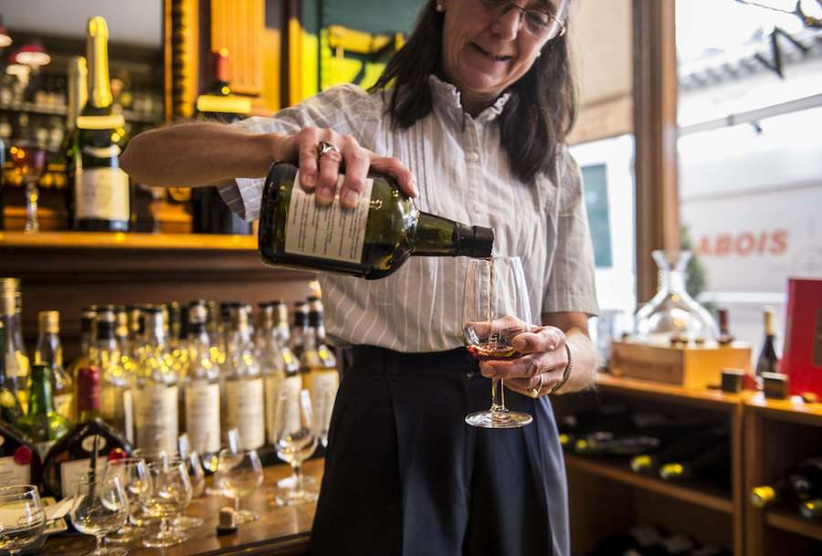 This image features a sommelier pouring a glass of wine. She is stood in a small wine store crowded with wine bottles.