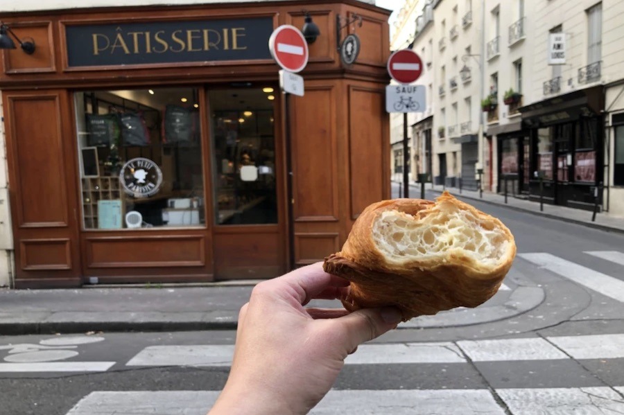 This is an image of a person holding up a half-eaten croissant in front of a patisserie.