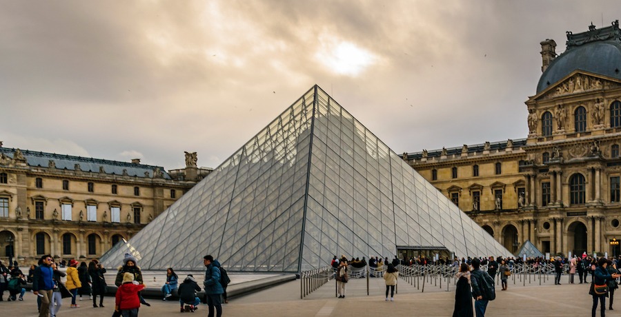 This is an image of the outside of the Louvre in Paris. The glass triangle entrance is in the centre of the image and people are gathered around it. The Louvre is one of the Top Guided Tours of Paris Attractions.