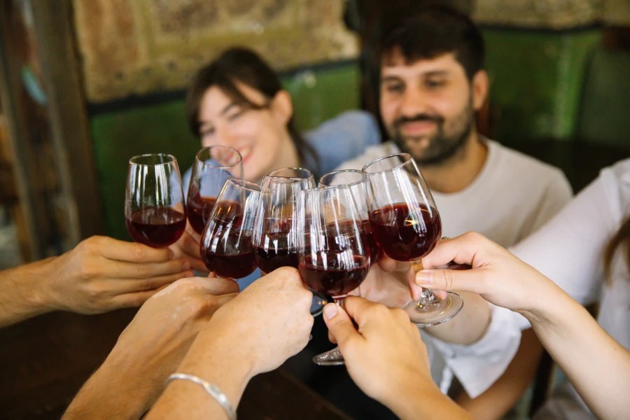 This is an image of a wine tasting. Several people are gathered round a table clinking wine glasses full of red wine.