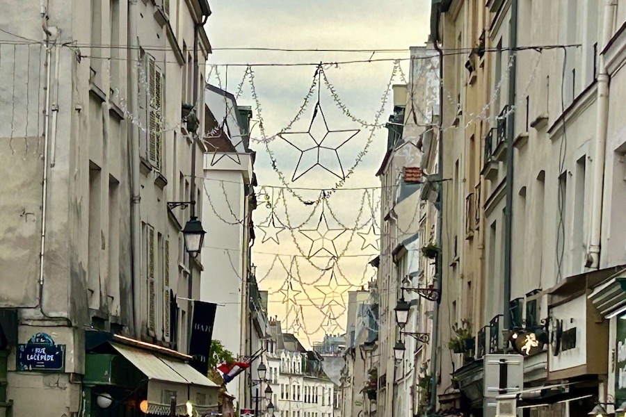 This is an image of a street in Paris with Christmas lights draped over the whole street from building to building.