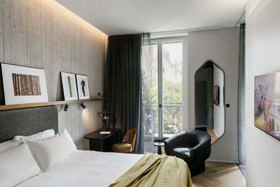 This is an image of a hotel room with a bed, a desk, a small chair and a mirror.