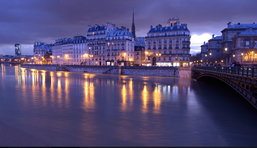 This is an image of a river in the evening. Lights from buildings in the city are reflecting on the surface of the water and it is gentle and pleasing.