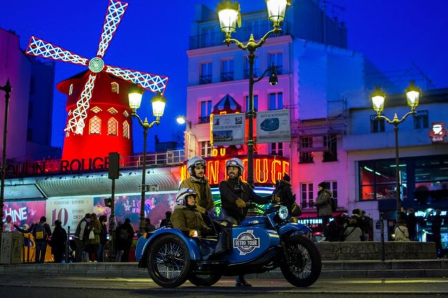 This is an image of Moulin Rouge at night. In front of it is a family of three riding in a side car.