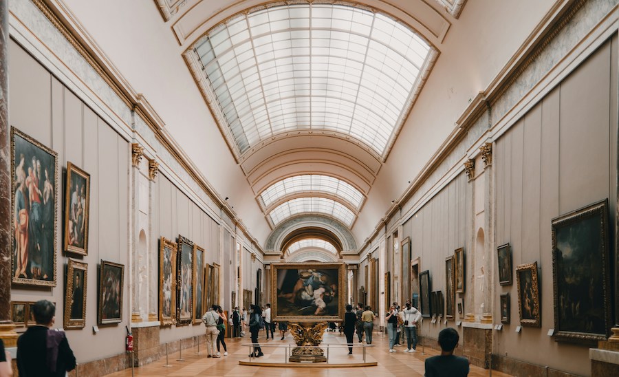 This is an image of one of the rooms in The Louvre. There are a bunch of people milling around viewing the art on the walls.