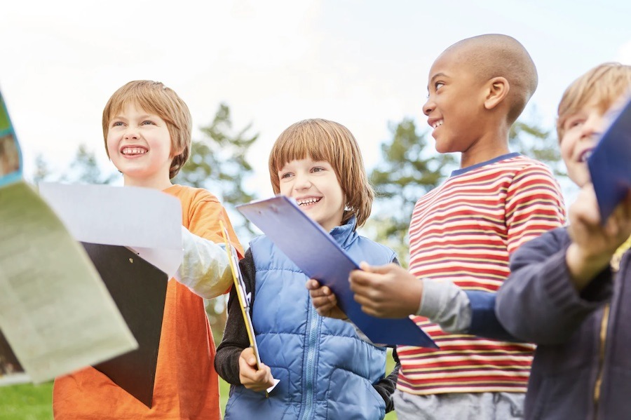This is an image of five kids holding clipboards, pens and papers ready to set off on a scavenger hunt. They are all smiling.