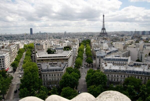 This is an image of a view from L'Arc De Triomphe that looks out on to the city of Paris. You can see the roundabout lanes and the tree-lined streets of Champs Elysees. You can also see the Eiffel Tower in the distance.