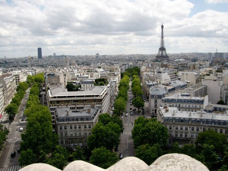 This is an image of a view from L'Arc De Triomphe that looks out on to the city of Paris. You can see the roundabout lanes and the tree-lined streets of Champs Elysees. You can also see the Eiffel Tower in the distance.