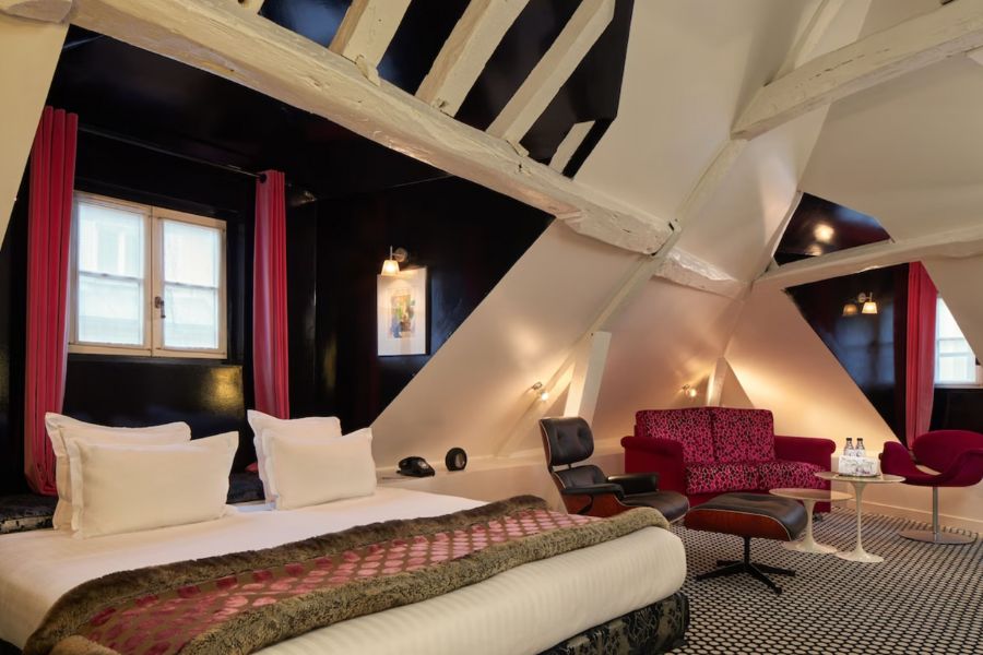 Cool Hotels In The Marais Paris: This is a large hotel room with a double bed. There is also a whole sitting area next to it with a red couch and coffee table.