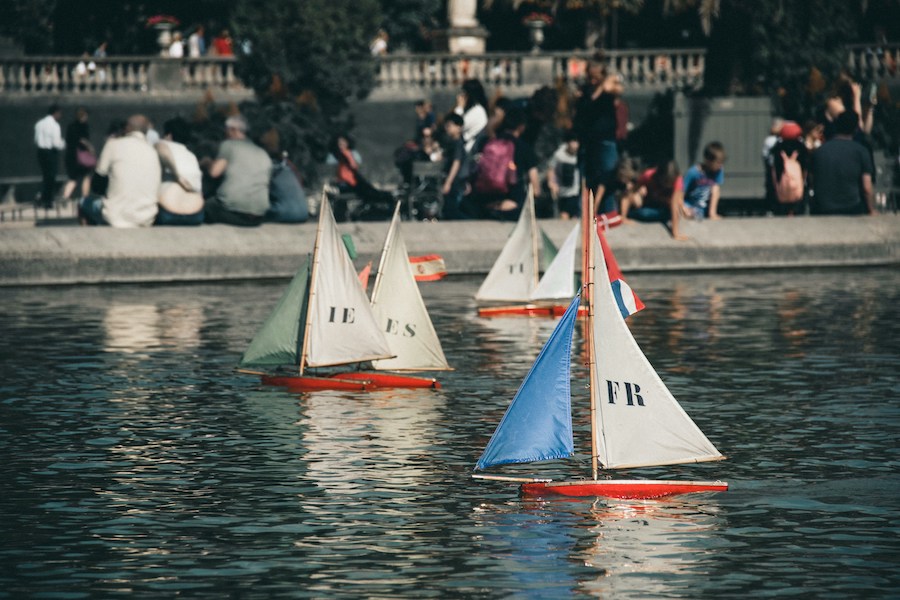This is an image of three toy boats floating around a small garden pond in Paris. There are people crowding in the background looking on.