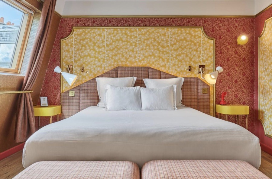 This is a picture of a stylish hotel room with yellow and pinkish-red accents. The double bed in the middle has cream covers.