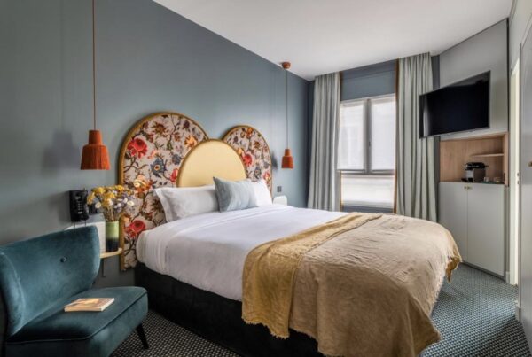 This is an image of a hotel room with blue painted walls. The bed is a double with a colourful headboard. There is a blue chair in the corner.