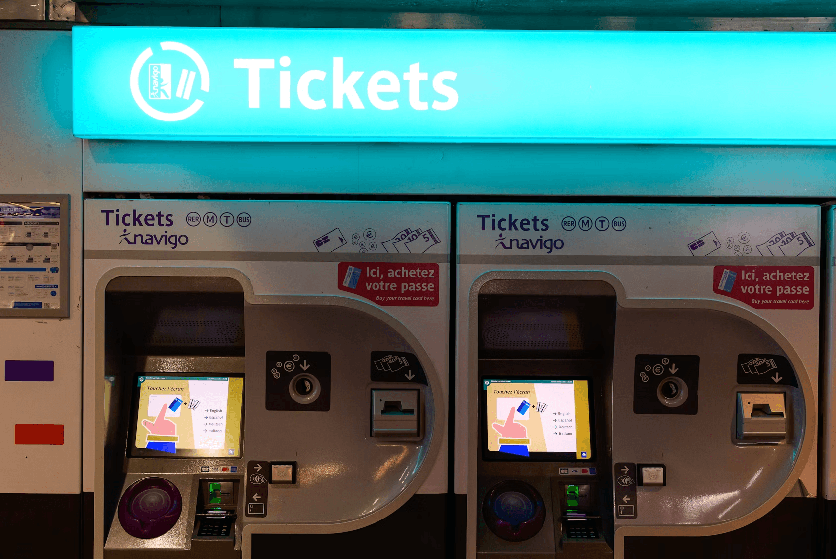 Two silver self-service ticket vending machines branded 'Tickets Navigo' in a Paris Metro station. Each machine has a touchscreen displaying multilingual options (English, Español, Deutsch, Italiano), coin and card payment slots, and a pink sticker reading 'Ici, achetez votre passe / Buy your travel card here.' The machines serve the RER, Metro, Tram, and Bus networks. Above them glows a large teal illuminated sign reading 'Tickets' with the Navigo logo.