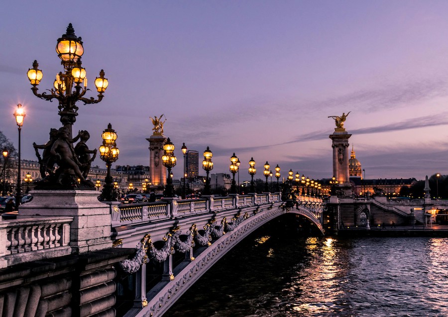 This is an image of Pont Alexandre which is a bridge in Paris that goes over the Seine River. It looks like sunset, as it is getting dark and the bridge street lights are on. An important thing to know before visiting Paris is that most places are safe, but it is important to still be cautious.