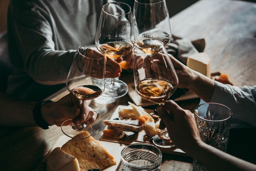 This is an image of a group of people clinking their wine glasses together. There is a cheeseboard in the middle of the table.