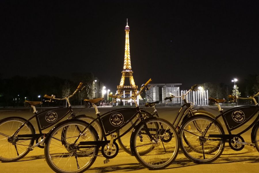 This is an image of three bikes propped up in front of a lit-up Eiffel Tower as it is the evening.