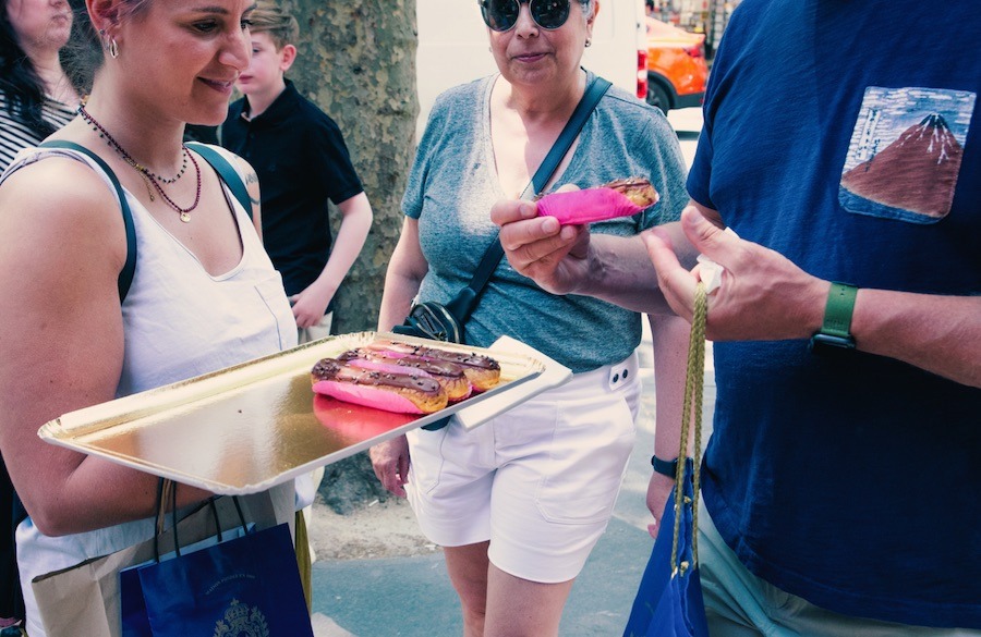 This is a image of a tour group crowded around a woman holding a tray of eclairs. It looks like they are all about to do a tasting.