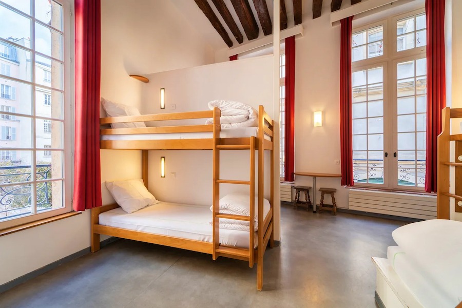 This is an image of a clean hostel bedroom with bunk beds with white bedding and long windows and high ceilings.