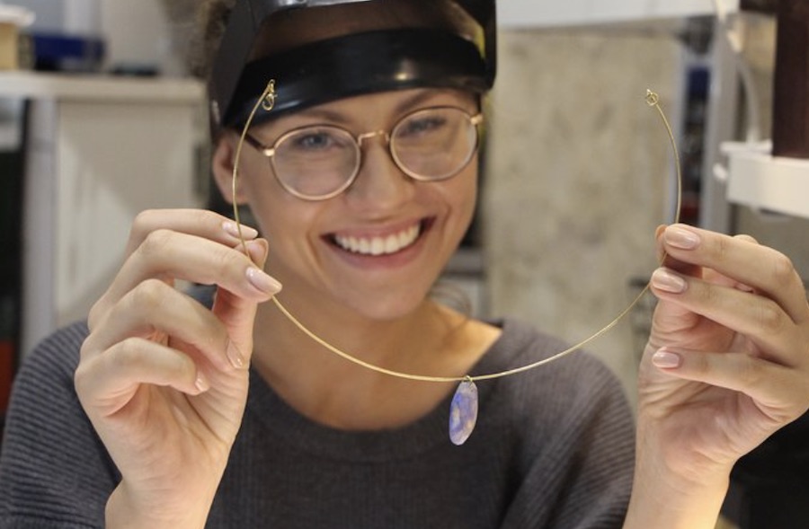 This is a picture of a lady wearing glasses, holding up a gemstone necklace that she looks to have just made.