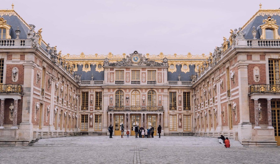 This is a picture of The Palace of Versailles. There are people milling around the front.