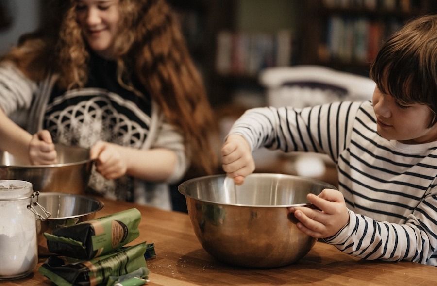 This is an image of two children baking. One is stirring ingredients in a mixing bowl. The other is adding ingredients to his bowl.